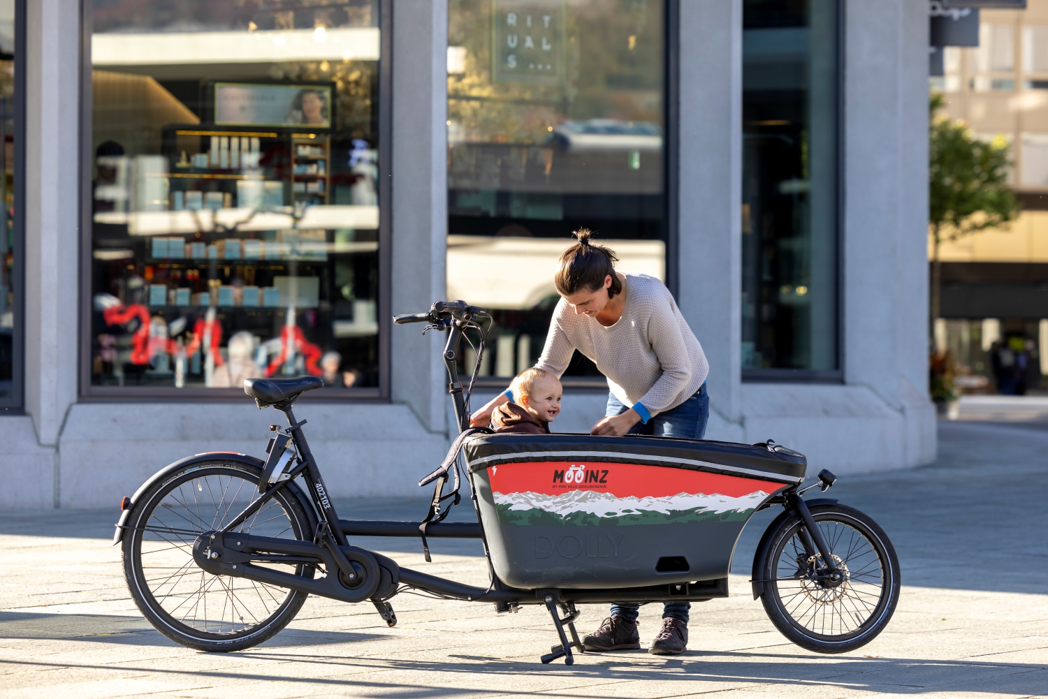 Junge Frau steht neben ihrem invia bikeshare Lastenrad, in welchem ihr kleines Kind sitzt.