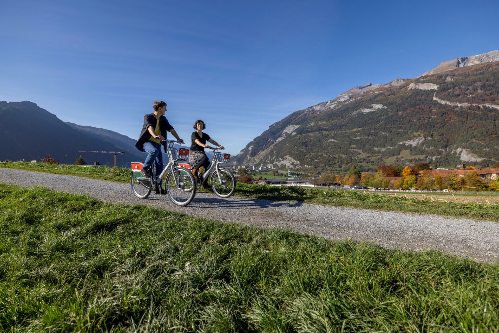 Frau und Mann fahren mit ihren invia bikeshare Rädern durch hügelige Landschaft, im Hintergrund die Berge